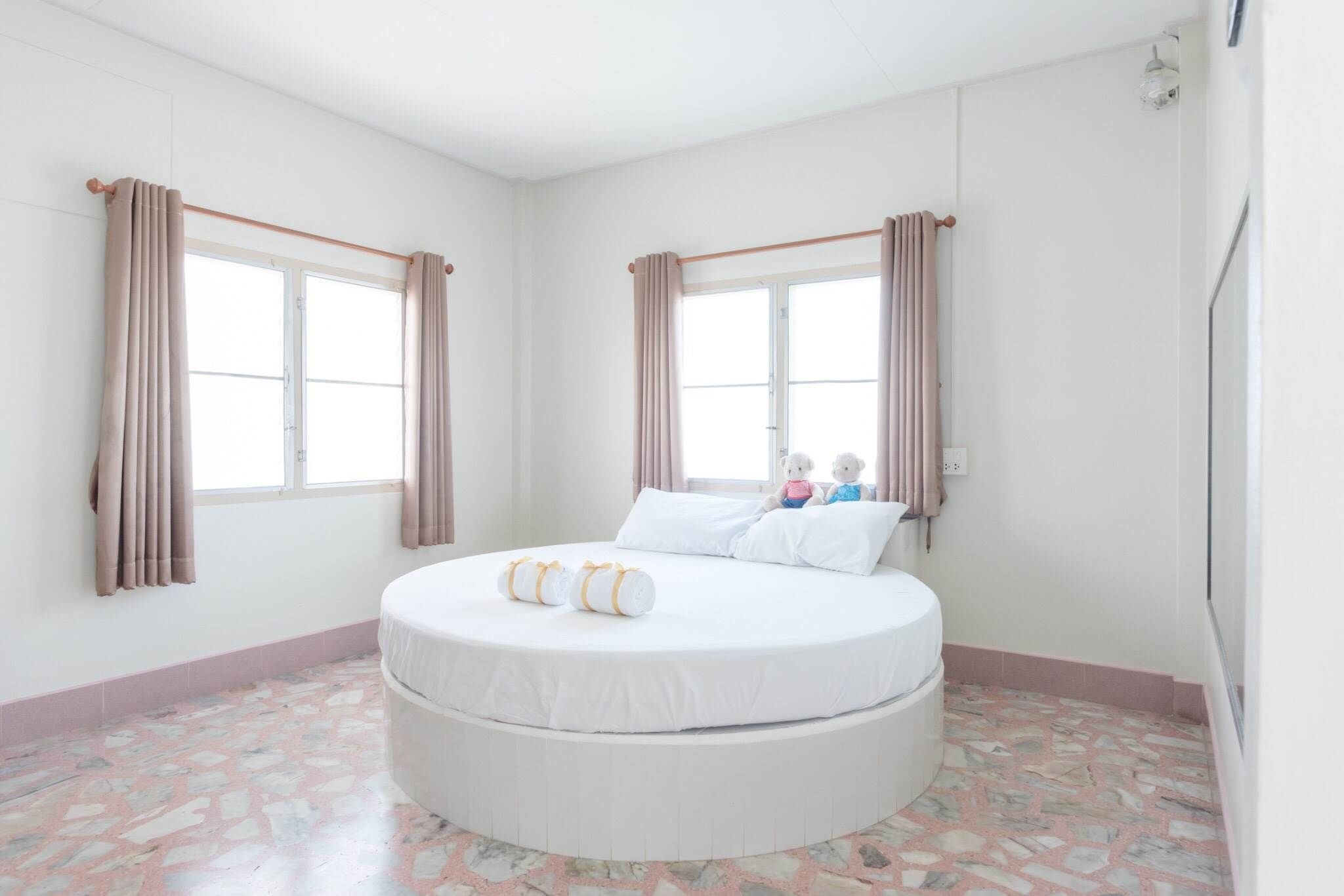 A circular bed in a room with two windows, a tiled floor, and curtains on the windows.