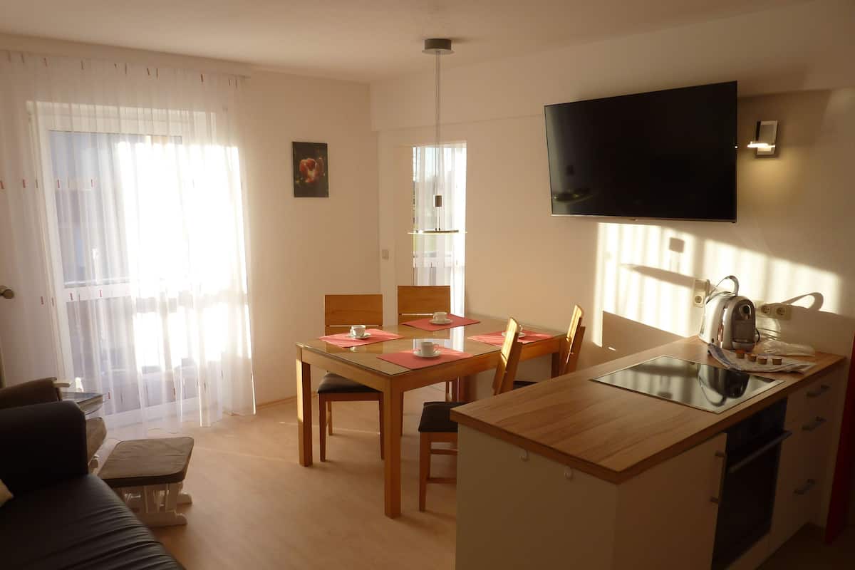 Una sala de estar moderna con un sofá, una mesa de comedor con sillas, una zona de cocina con un televisor y una ventana con cortinas.