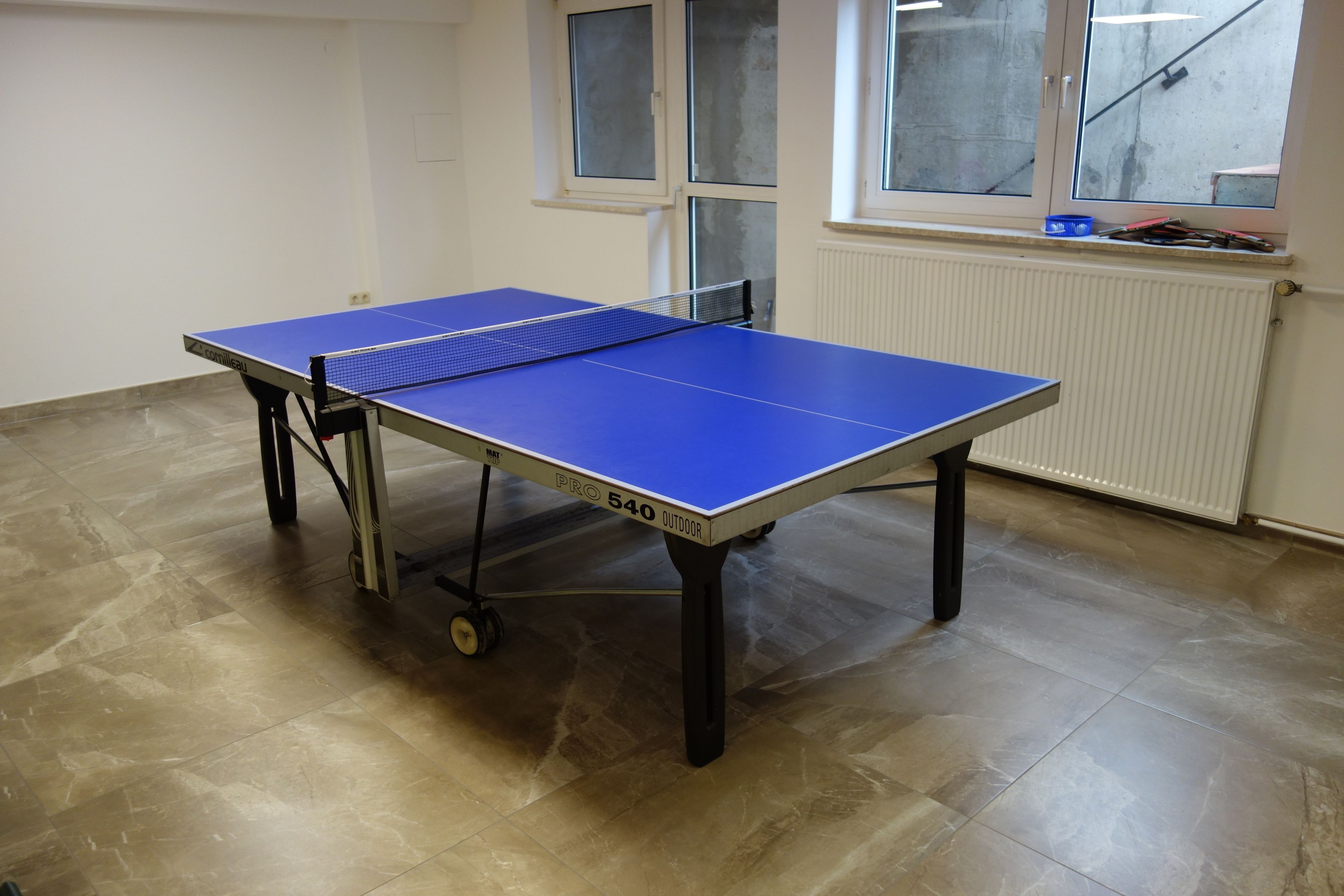 A table tennis table with wheels, positioned in a room with tiled flooring and a window.