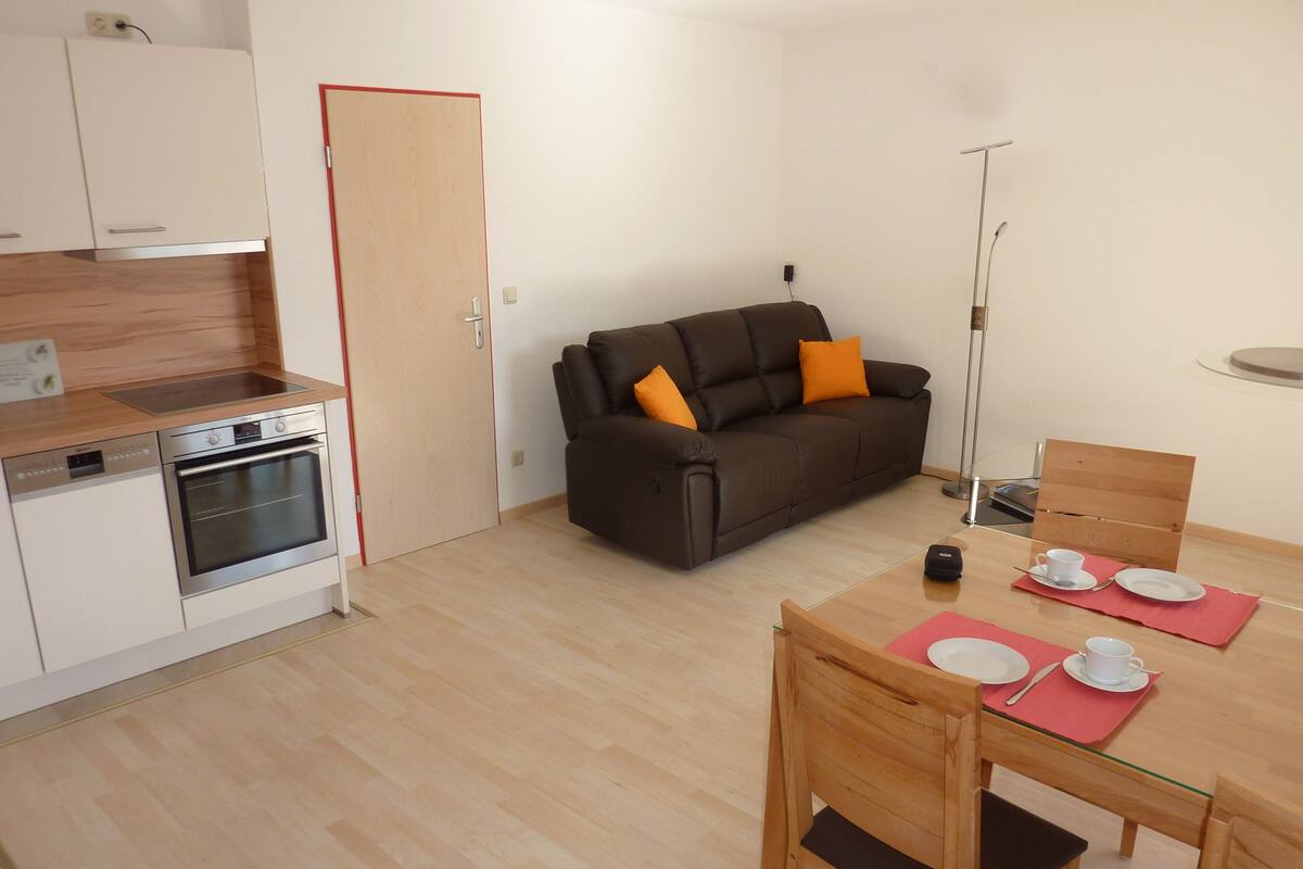 Un espacio habitable compacto con kitchenette, sofá, mesa de comedor con sillas y una lámpara de pie.