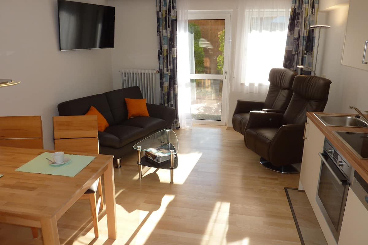 Una sala moderna con un sofá, una mesa de comedor, una mesa de centro, un televisor montado en la pared y una cocina con un lavamanos y un horno.