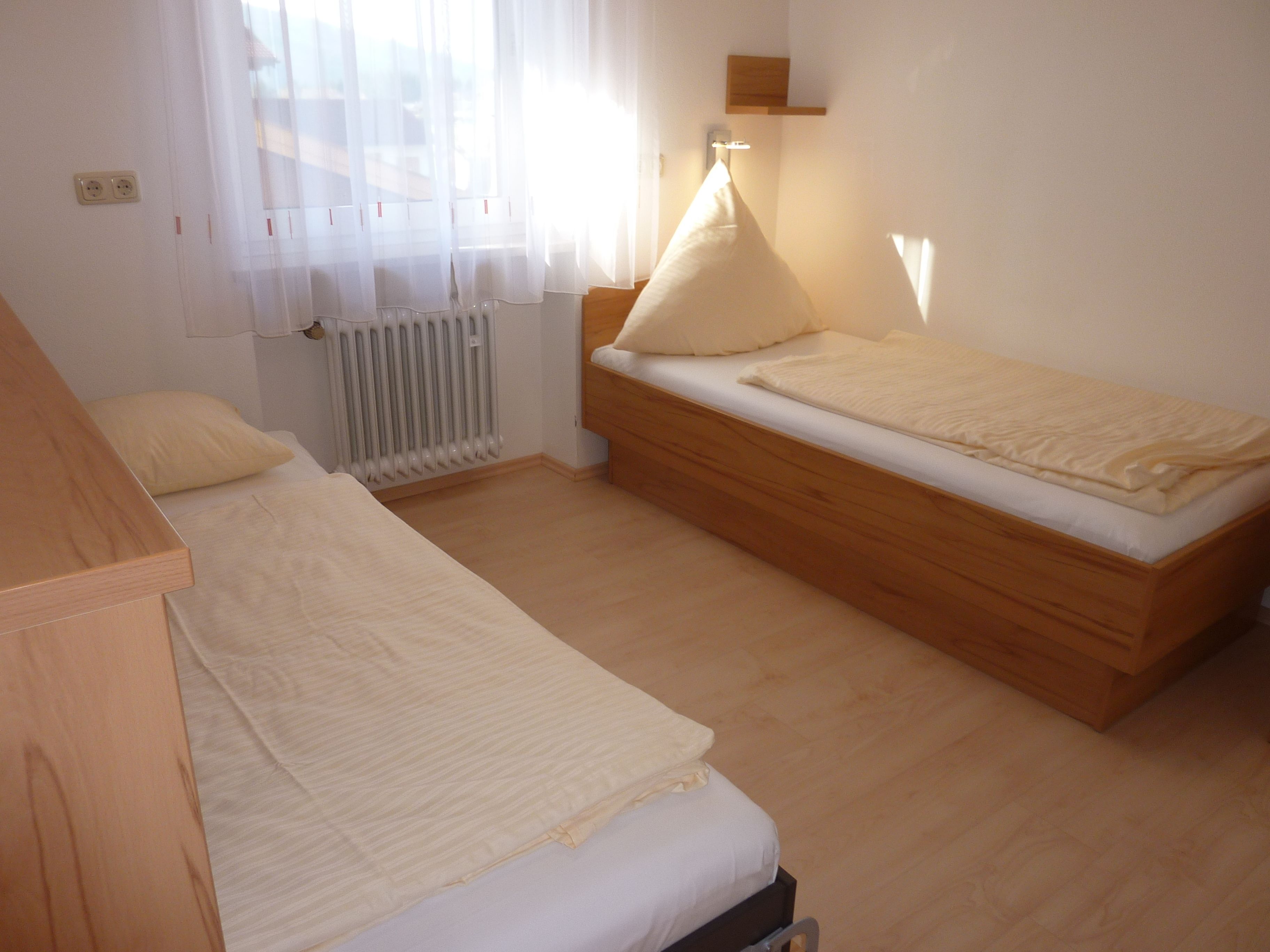 Een slaapkamer met twee eenpersoonsbedden, een radiator en een raam met dunne gordijnen.