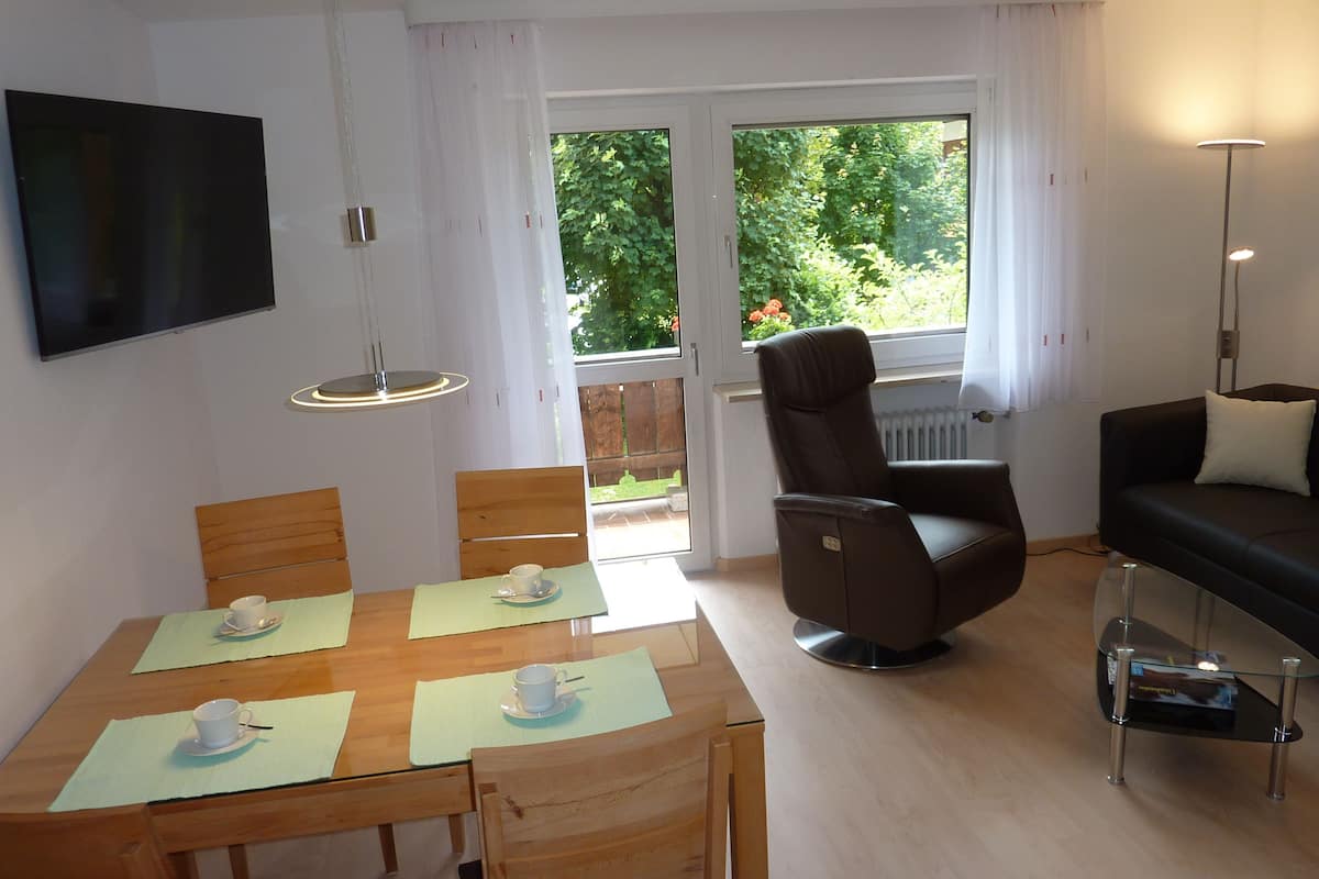 Una sala de estar moderna con comedor, un sofá, un televisor y vista a áreas verdes.