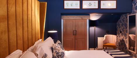A bedroom with a large bed, wooden headboard, and a wardrobe. There are framed pictures on the wall and a chair in the corner.