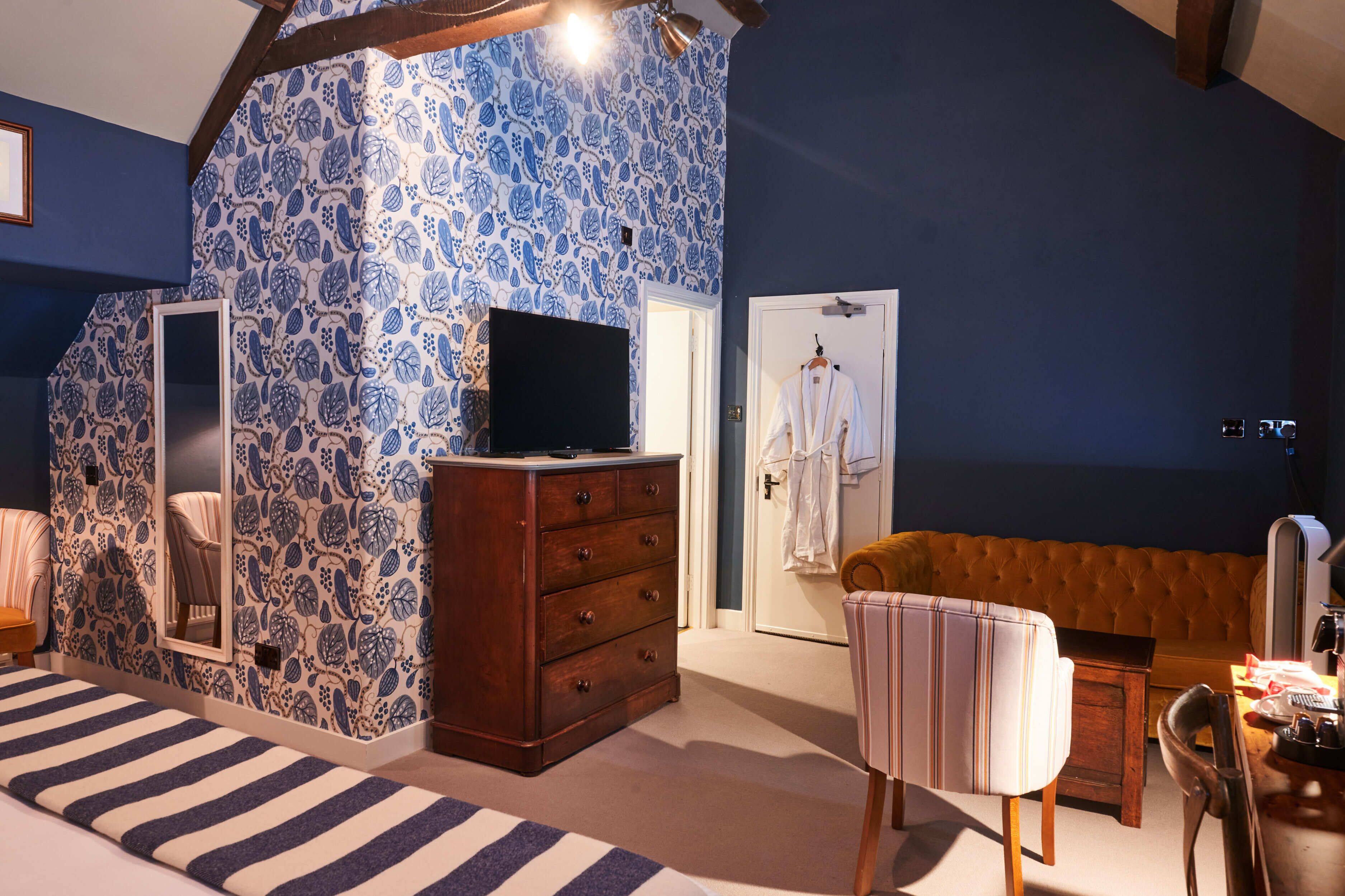 A bedroom with a patterned wall, a wooden dresser, a television, a bed with striped bedding, a chair, and a sofa.