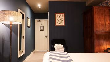 Ein Hotelzimmer mit Bett, Sessel, Tür, Spiegel und einer Wand mit einem gerahmten Pflanzenbild.