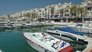 A marina with multiple boats docked, surrounded by buildings with balconies and palm trees.