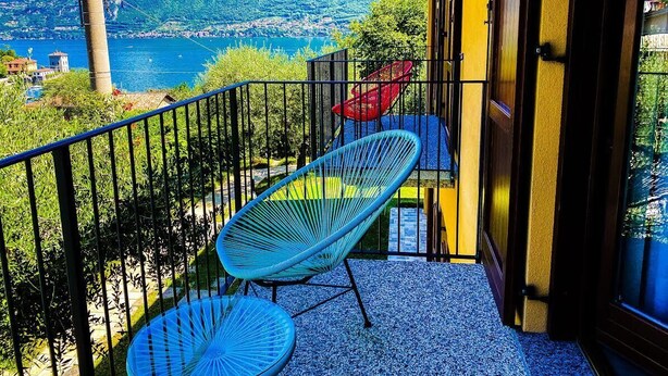 Un balcón con vista a un lago y montañas, una silla azul y una silla roja.