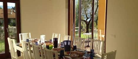 Comedor con una mesa dispuesta para cuatro personas, sillas blancas, una araña y un amplio ventanal con vistas a la vegetación.