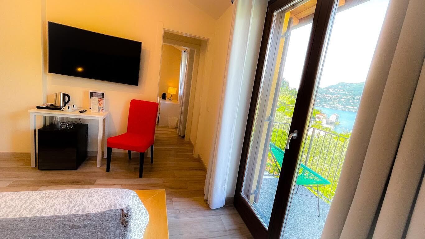 Habitación de hotel con televisor, una silla roja, una mesita con tetera y vistas a un balcón con mesa y sillas.