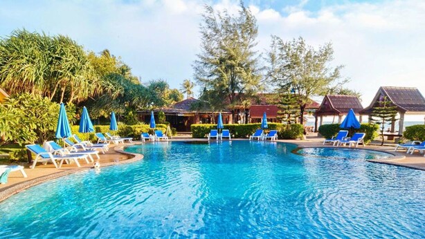 A swimming pool with lounge chairs and umbrellas, surrounded by greenery and buildings. - Prukrom Guesthouse (Ko Lanta)
