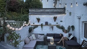 Un rincón acogedor al aire libre con pared de piedra, luces colgantes y plantas en macetas.