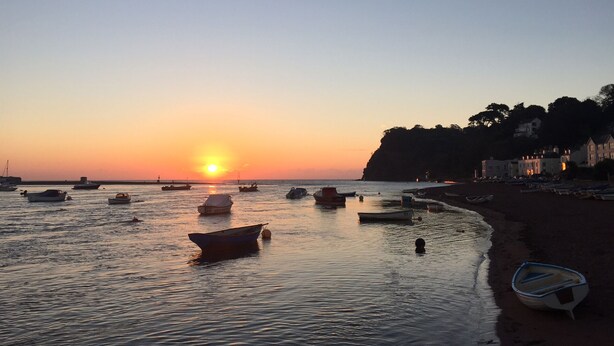 Escena de playa con barcos, una puesta de sol y edificios a la orilla.