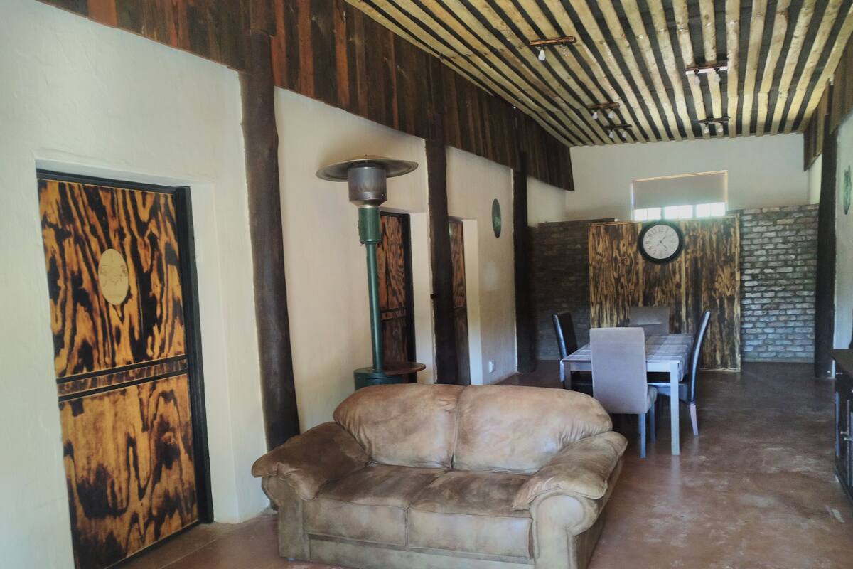 A living room with a sofa, a dining table with chairs, a clock on the wall, and a wooden ceiling.