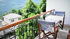 Un balcon avec une balustrade en bois, une table recouverte d’une nappe blanche, et une vue sur des collines verdoyantes et un bâtiment blanc.
