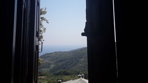 On aperçoit, à travers une fenêtre ouverte, un bâtiment dont le toit est recouvert de tuiles, un ciel dégagé et, au loin, un paysage vallonné.