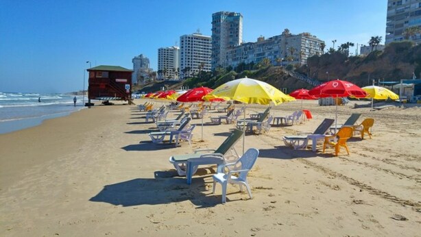Una playa con filas de tumbonas y sombrillas, una torre de salvavidas y edificios altos al fondo.