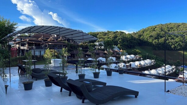 An outdoor seating area with lounge chairs and potted plants, a covered structure, and a view of a hillside with parked vehicles. - Mon Ing Dao (Mae Rim)