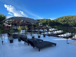 Zona de estar al aire libre con sillones reclinables y plantas en macetas, una estructura cubierta y vistas a una ladera con vehículos estacionados.