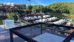 A rooftop terrace with a metal grid floor, a large white water feature, and a view of a residential area with houses and green hills. - Mon Ing Dao (Mae Rim)