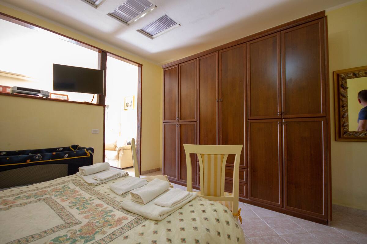 A bedroom with a bed, a wooden wardrobe, a television on a stand, and a chair.