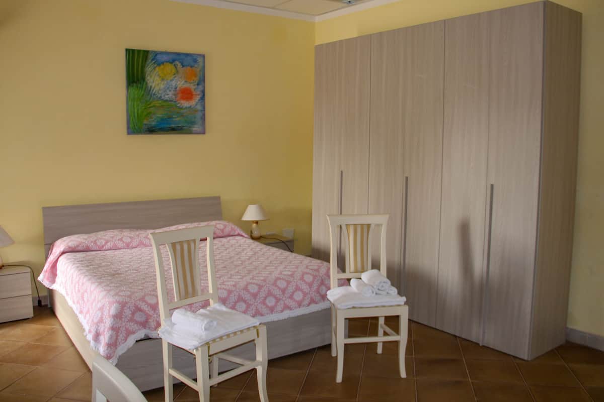 A bedroom with a bed, two chairs, a wardrobe, and a painting on the wall.