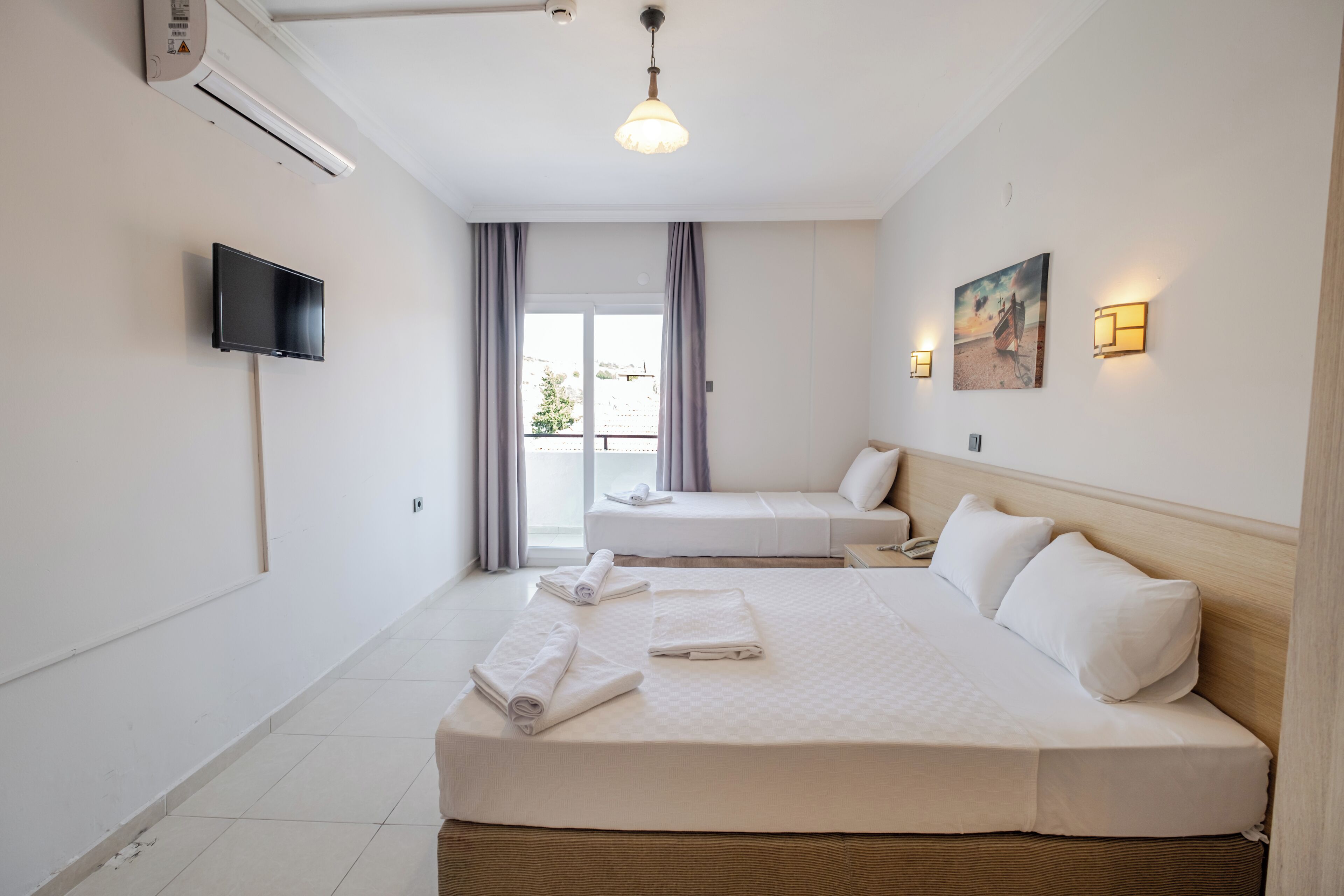 Quarto de hotel com uma cama grande, uma cama menor, uma TV de tela plana, um ventilador de teto e uma janela com cortinas.