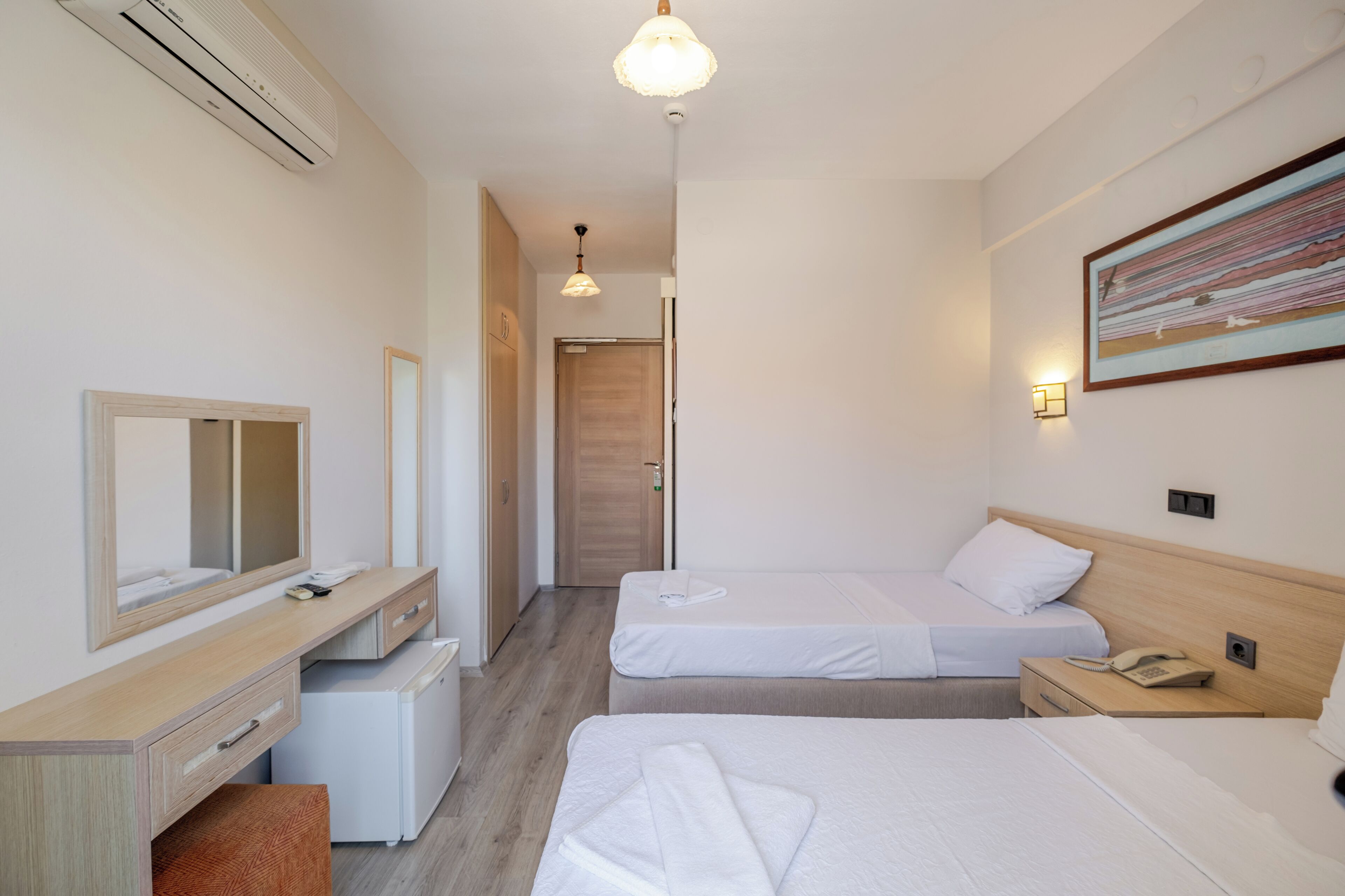 Habitación de hotel con dos camas, un escritorio con espejo, un refrigerador pequeño y un cuadro en la pared.