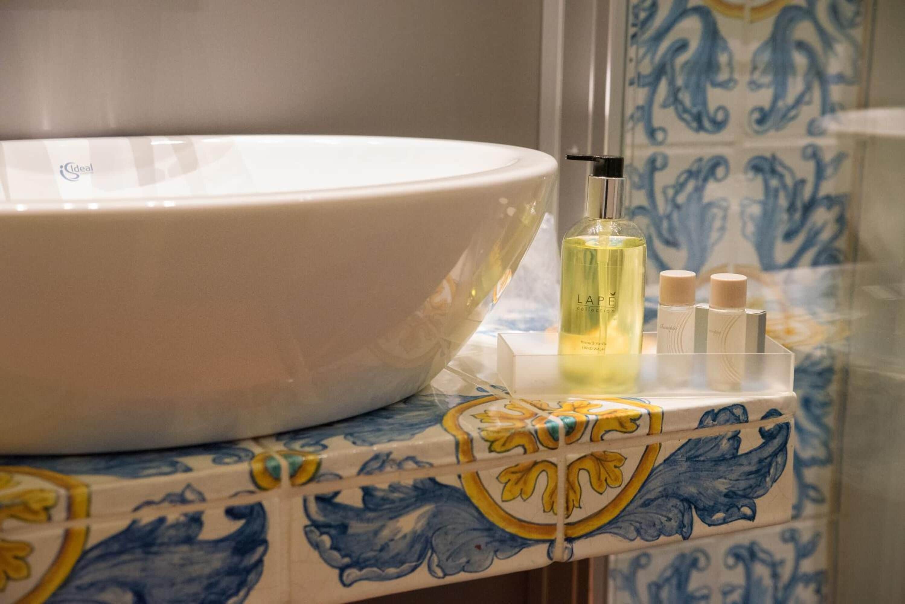 Un baño con un lavamanos blanco, una botella de loción y un dispensador de jabón sobre una encimera con baldosas.