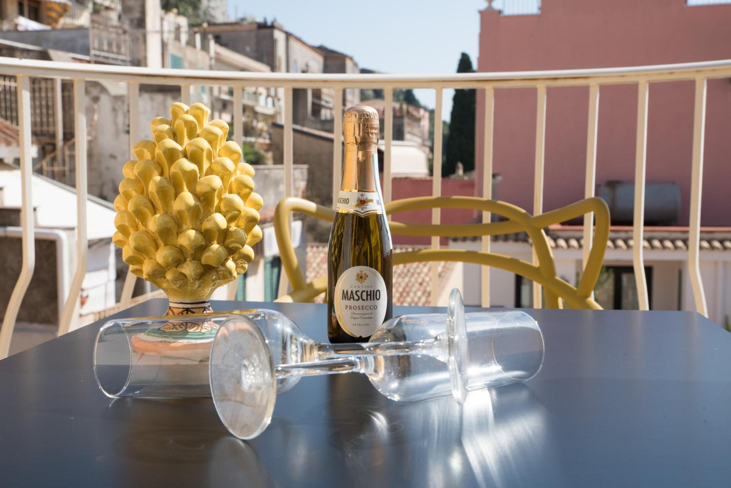 Una mesa con una botella de Maschio Prosecco, dos copas de vino y una piña dorada decorativa en un balcón con barandilla y edificios al fondo.