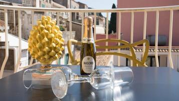Una mesa con una botella de Maschio Prosecco, dos copas de vino y una piña dorada decorativa en un balcón con barandilla y edificios al fondo.
