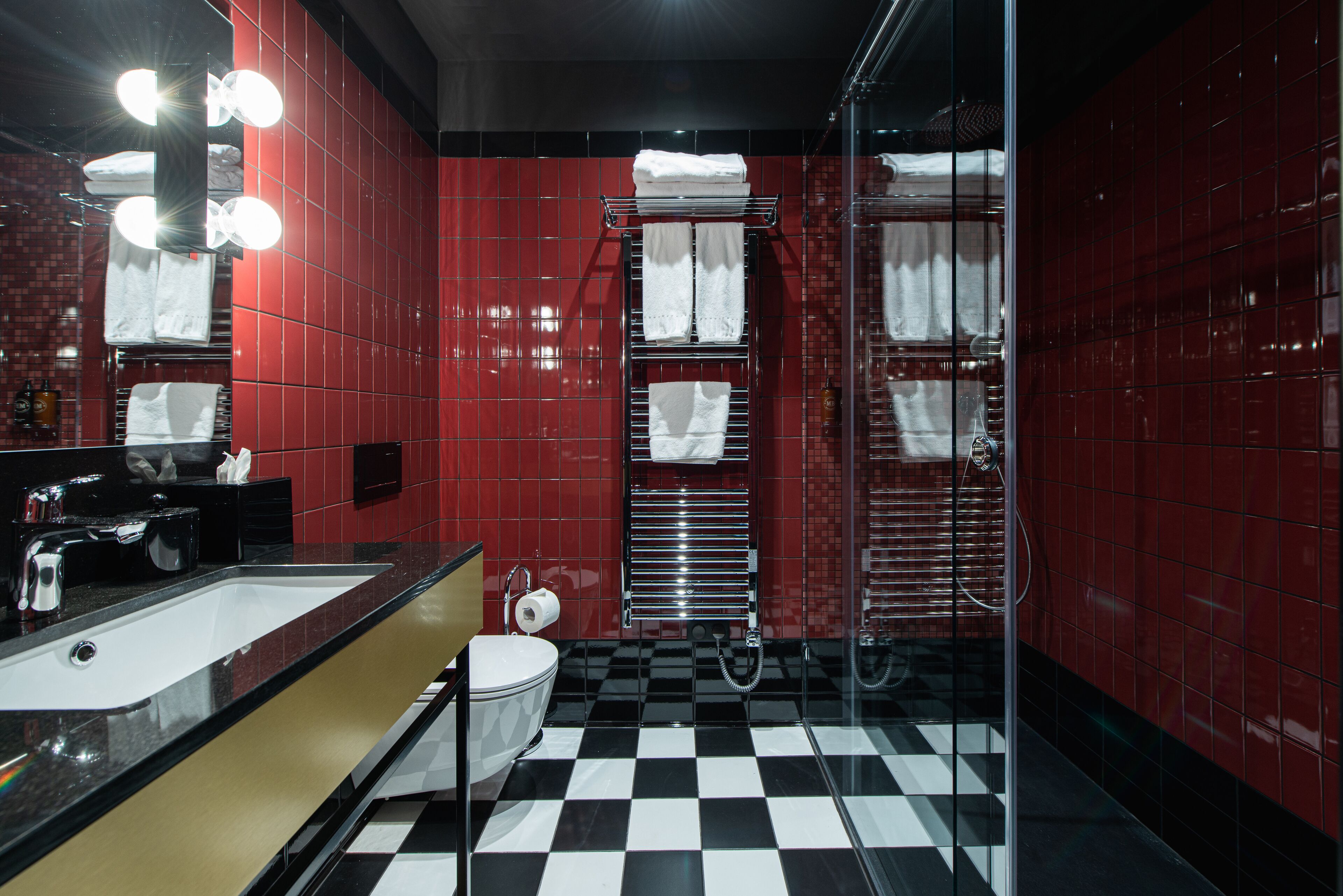 Une salle de bain avec un mur recouvert de carreaux rouges, un sol à carreaux noirs et blancs, un lavabo, des toilettes et une cabine de douche en verre.