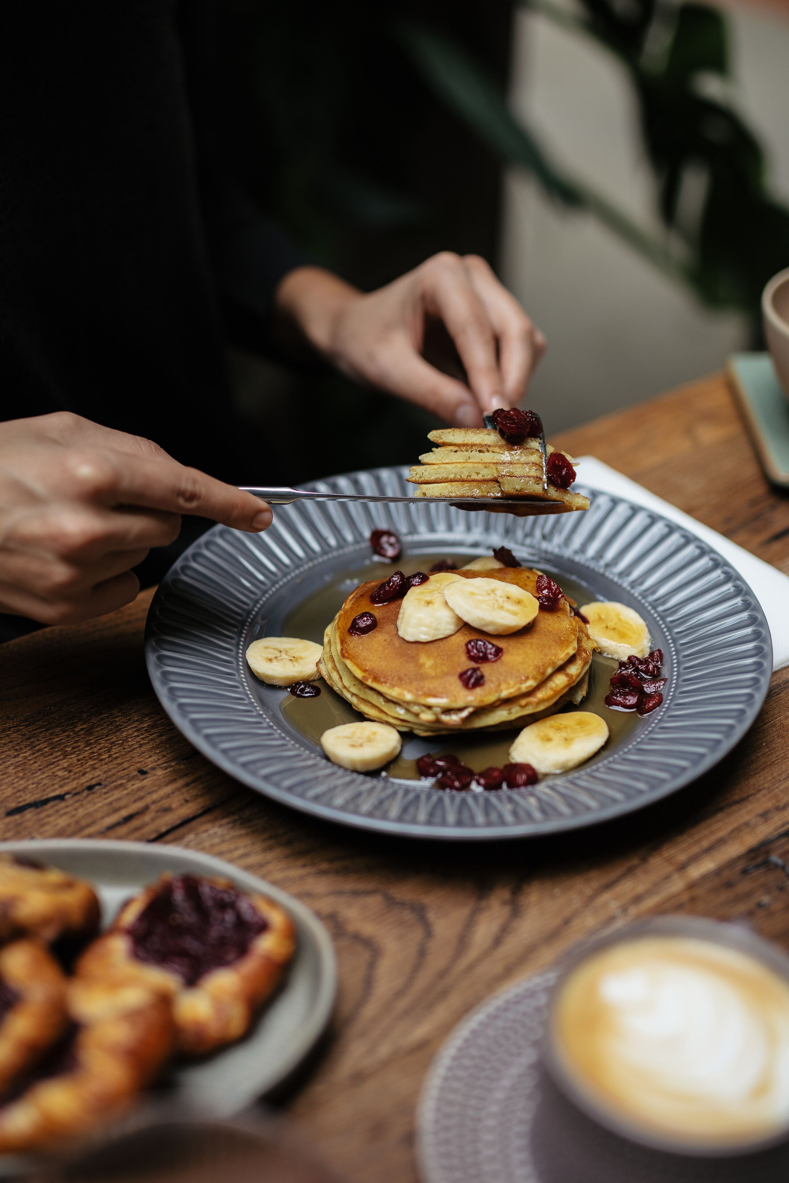 A plate of pancakes with bananas and berries, a cup of coffee, and a plate of pastries on a wooden table.