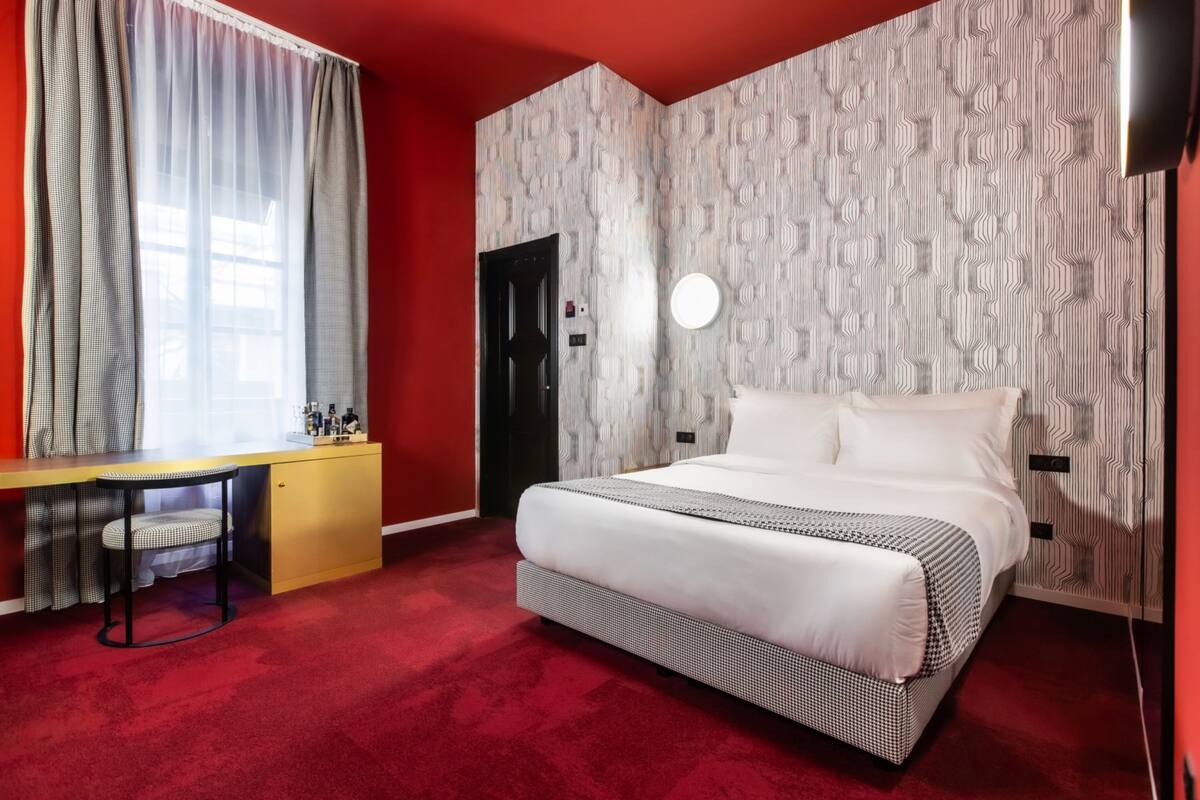 Une chambre d’hôtel avec un grand lit, un bureau avec une chaise, une armoire et un sol recouvert d’une moquette rouge.
