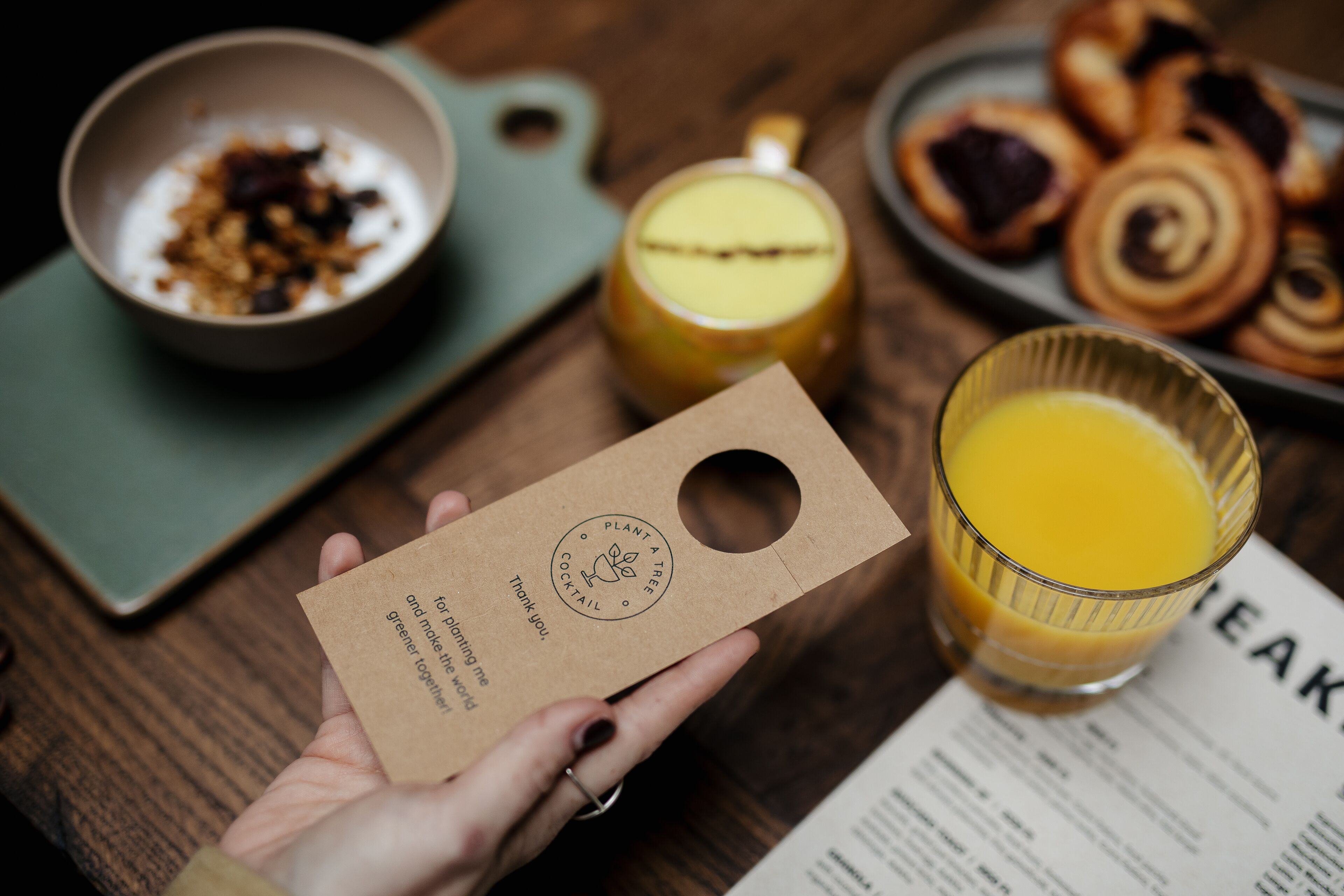A hand holding a brown paper bag with a logo and text, a glass of orange juice, a bowl of granola, a plate of pastries, and a menu on a wooden table.