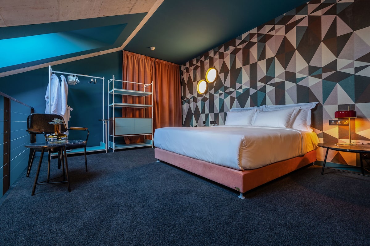 Une chambre d’hôtel moderne, équipée d’un lit, d’un bureau, d’une chaise, d’un porte-manteau et d’un mur orné d’un motif géométrique.