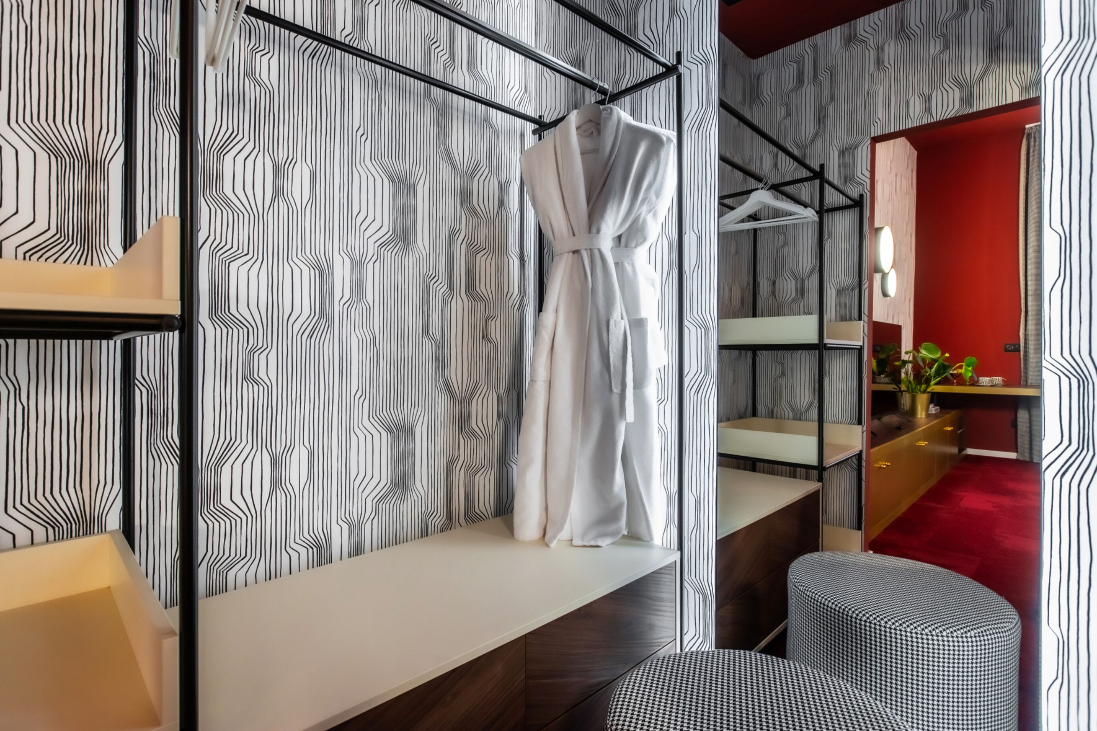 A modern bathroom with a striped wallpaper, a white robe hanging on a rack, a wooden vanity, and a red accent wall.