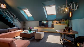 A modern living room with a sofa, coffee table, and a dining area with a table and chairs. There is a TV on a stand, a staircase, and a skylight.