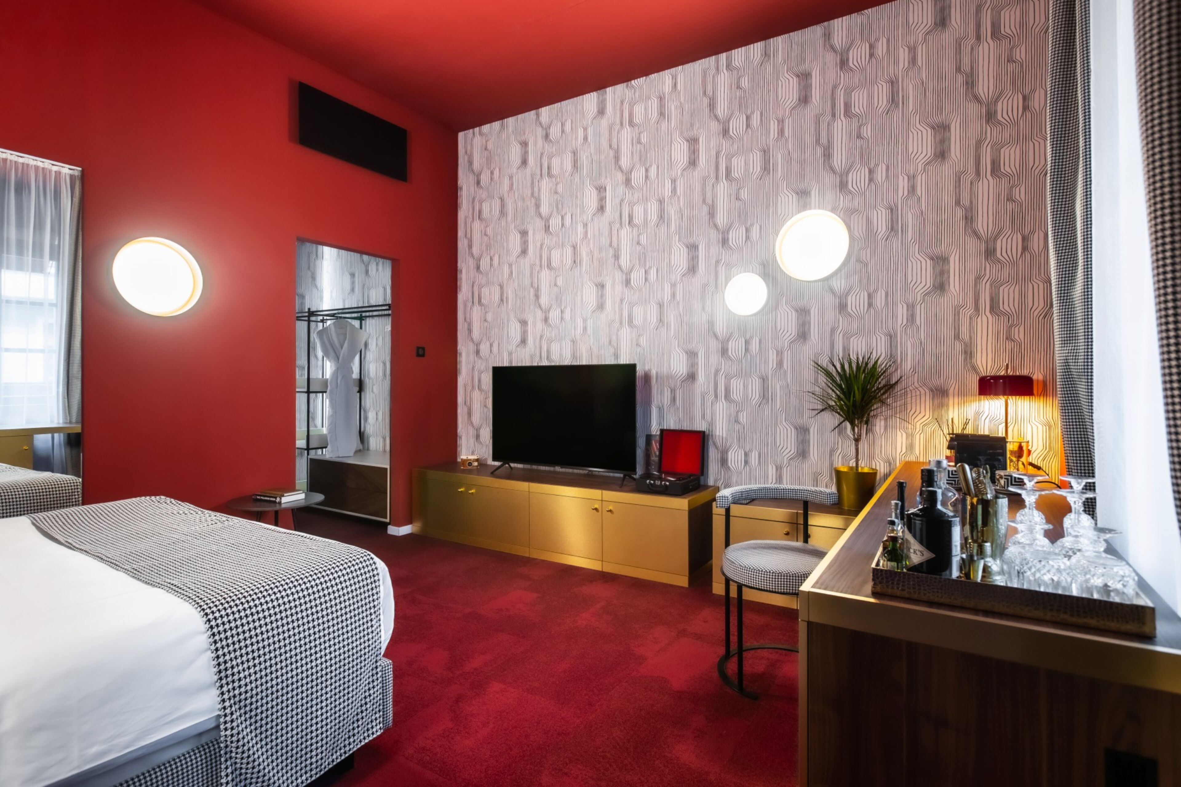 Ein Hotelzimmer mit rotem Teppich, einem Fernseher auf einer goldenen Kommode, einer Bar mit Flaschen und einer Wand mit Muster-Tapete.
