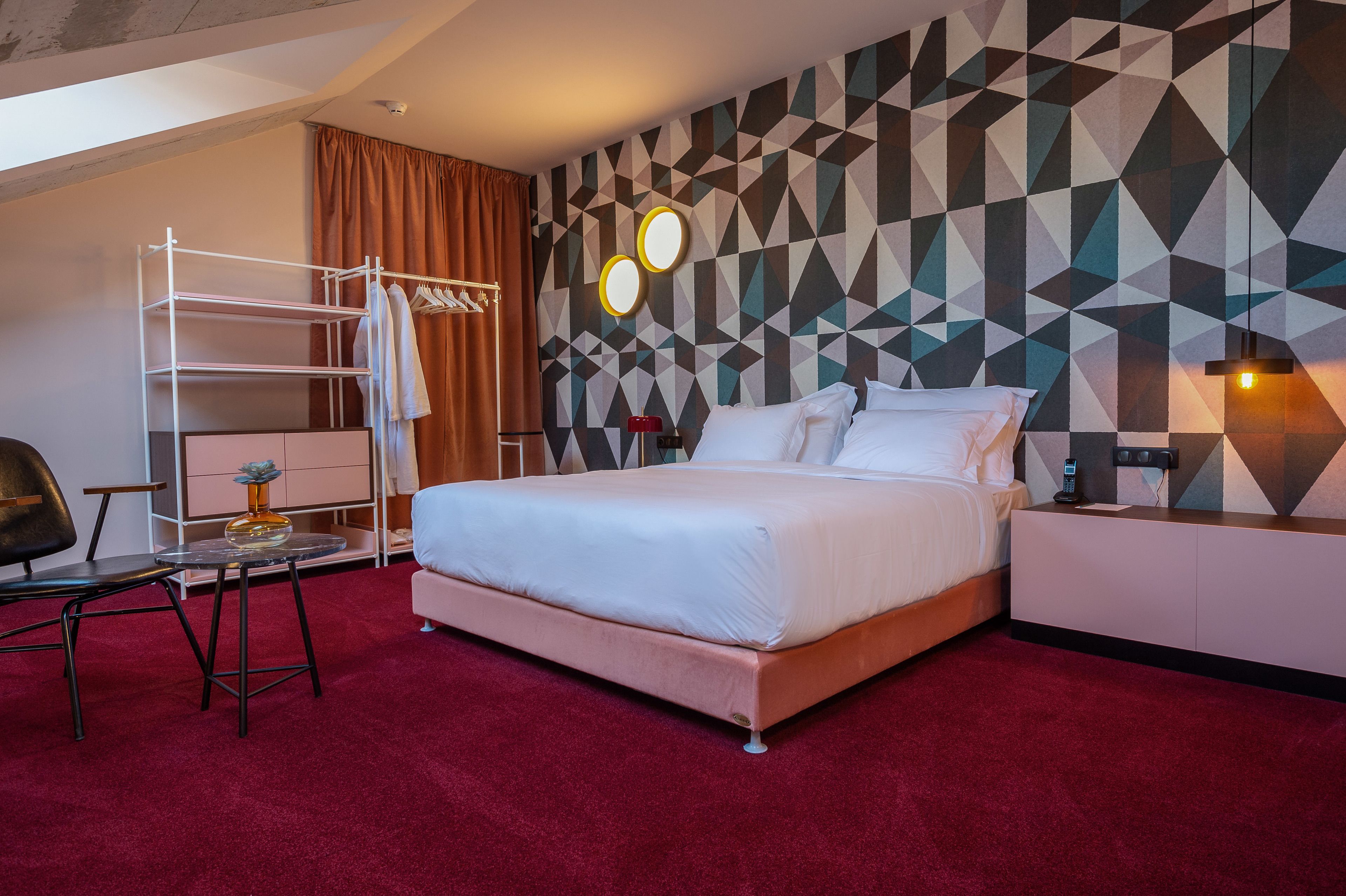 A hotel room with a bed, a desk with a chair, a coat rack, and a wall with geometric patterns.