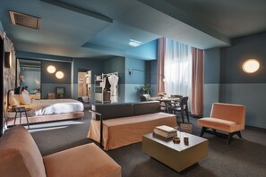 Una habitación de hotel moderna con cama, sofá, sillón y un rincón de comedor con mesa y sillas.