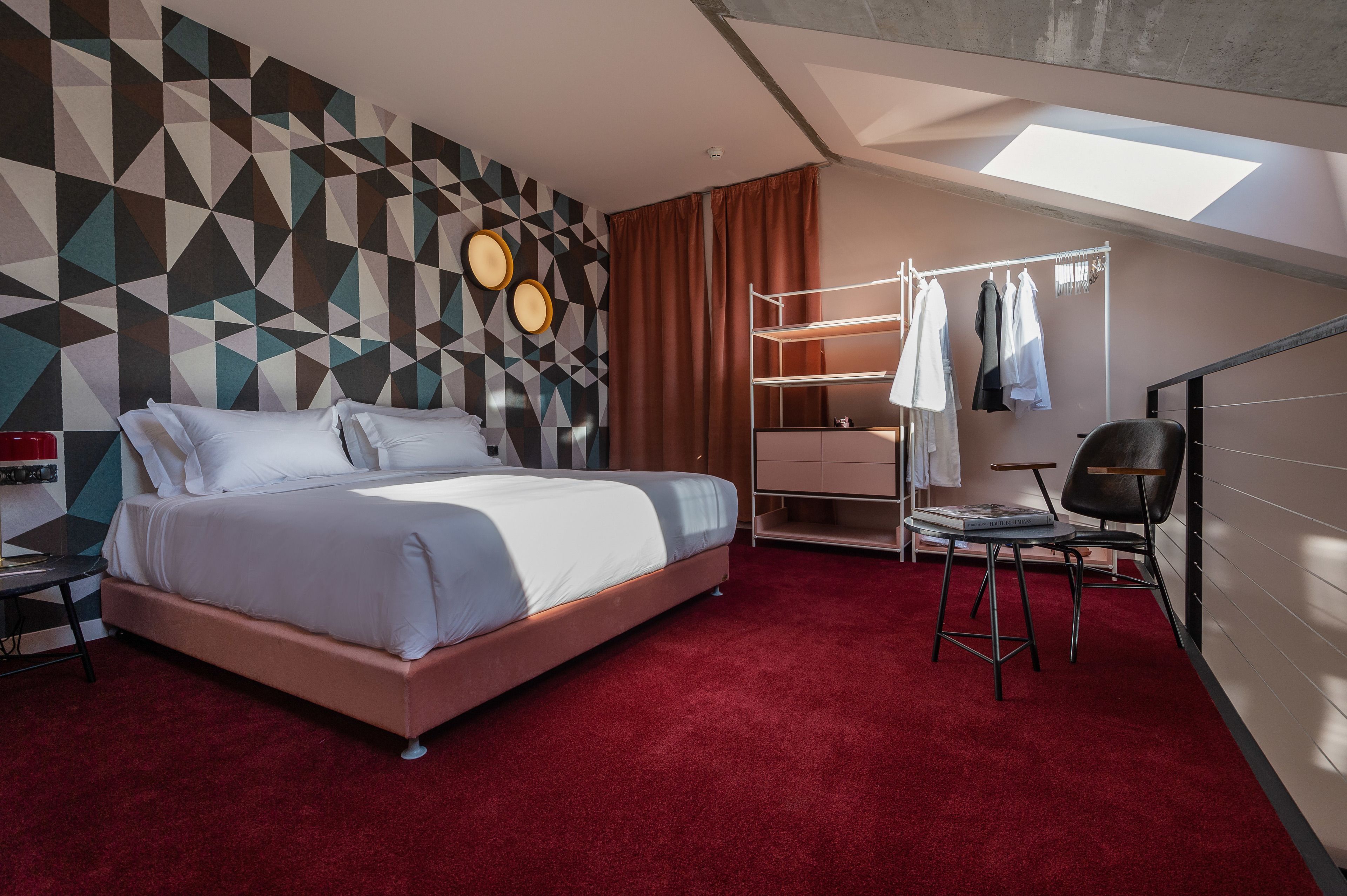 A modern bedroom with a geometric-patterned wall, a bed with white linens, a red carpet, a clothing rack, and a skylight.