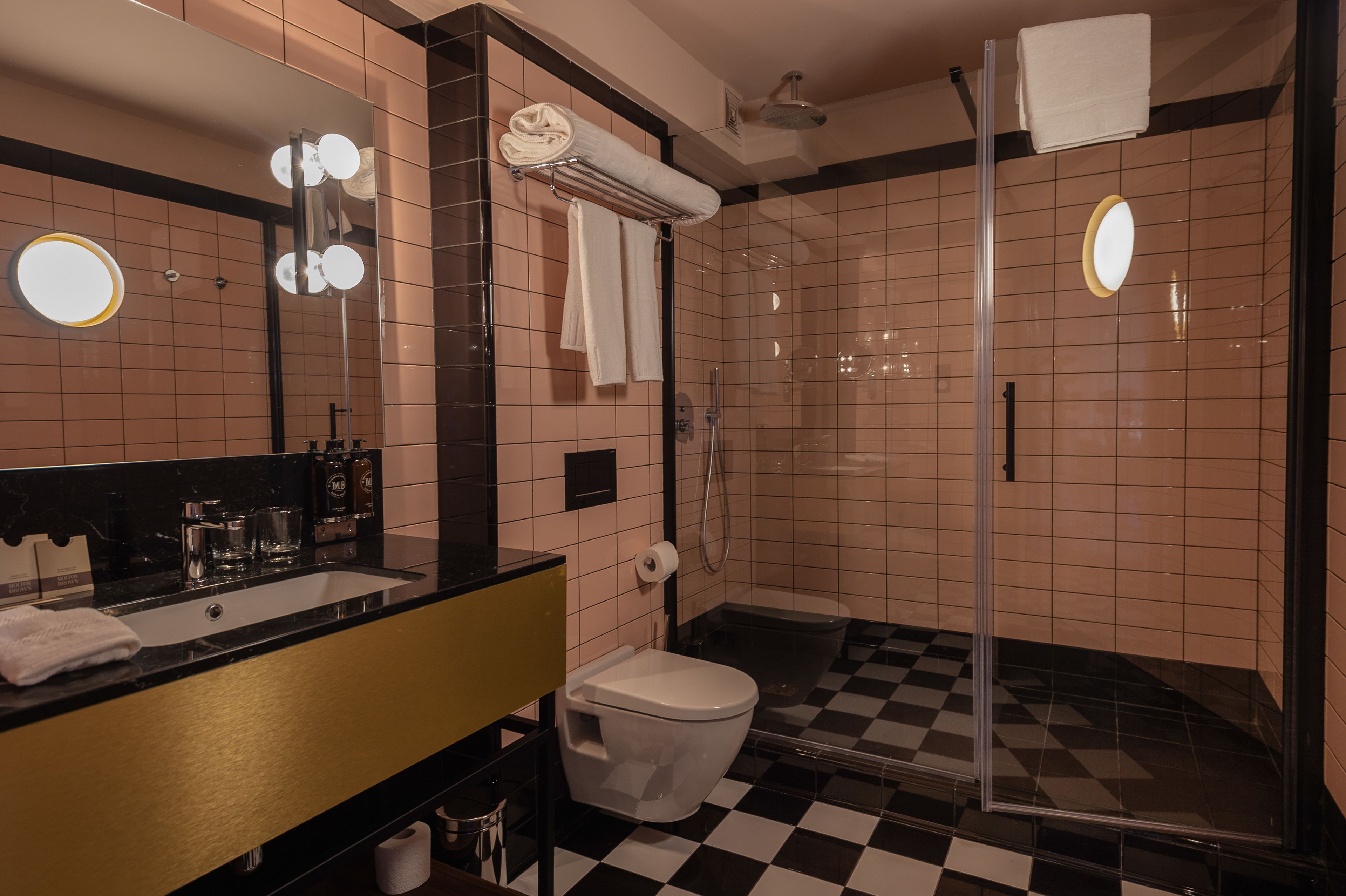 A bathroom with a black and white checkered floor, a white toilet, a glass-enclosed shower, a mirror with lights, and a towel rack with towels.