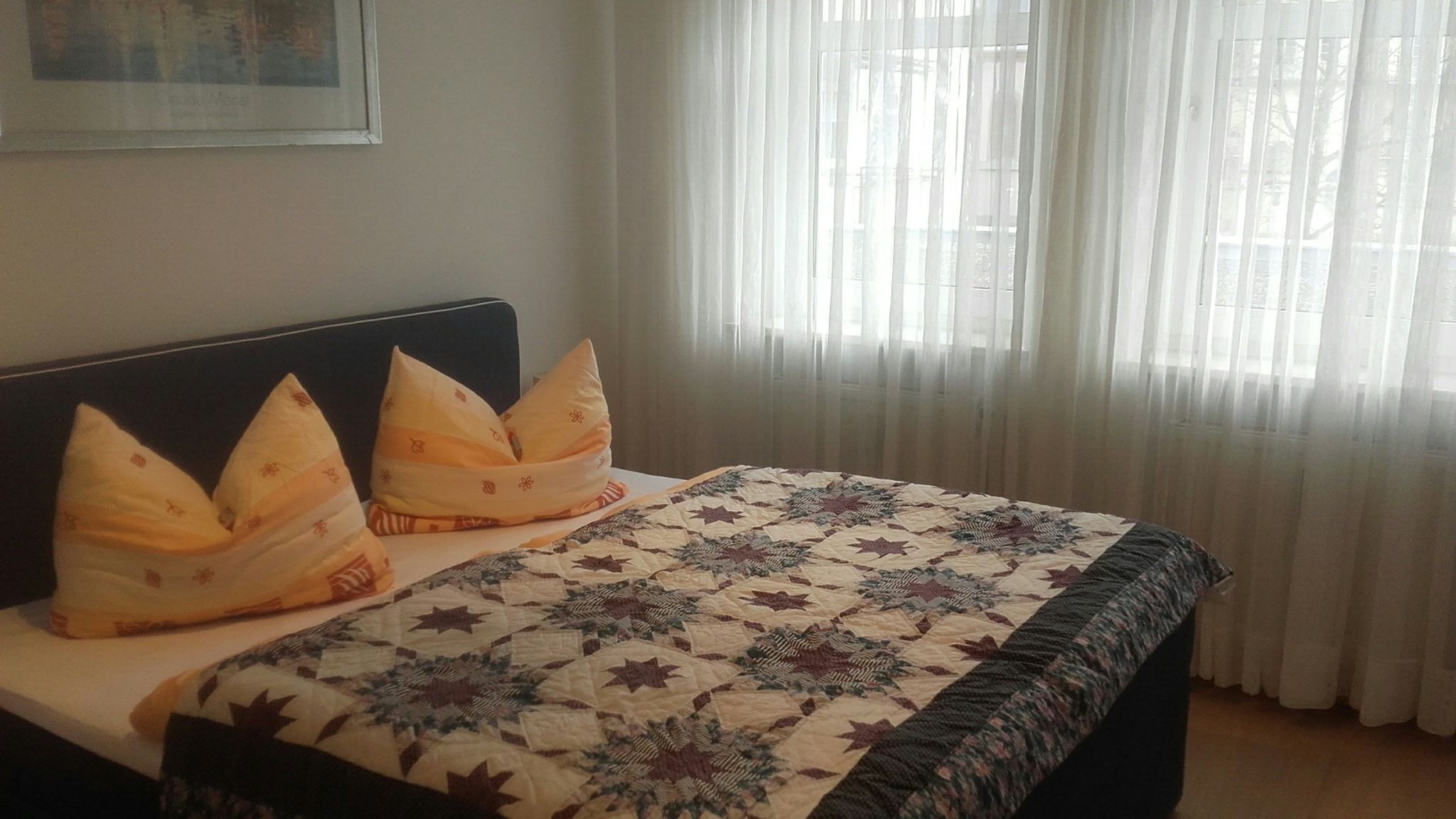 A neatly made bed with a patterned quilt and two decorative pillows, a window with sheer curtains, and a framed picture on the wall.