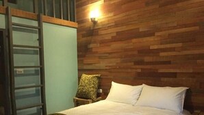 Une chambre à coucher avec une tête de lit en bois, un lit recouvert de draps blancs, un plaid vert et une chaise agrémentée d’un coussin à motifs.