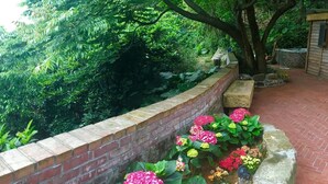 Un chemin pavé de briques, bordé d’un côté par un mur de briques et de l’autre par un jardin luxuriant, où s’épanouissent différentes espèces de fleurs et où se dresse un arbre.