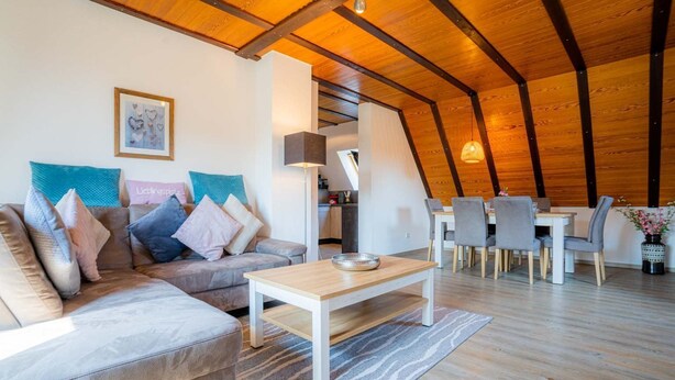 A modern loft-style living space with a wooden ceiling, a sofa with cushions, a dining table with chairs, and a kitchen area in the background. - Apartment 5 "Bellmann" (ID 024) - House Dorfstraße 30a (Sankt Peter-Ording)