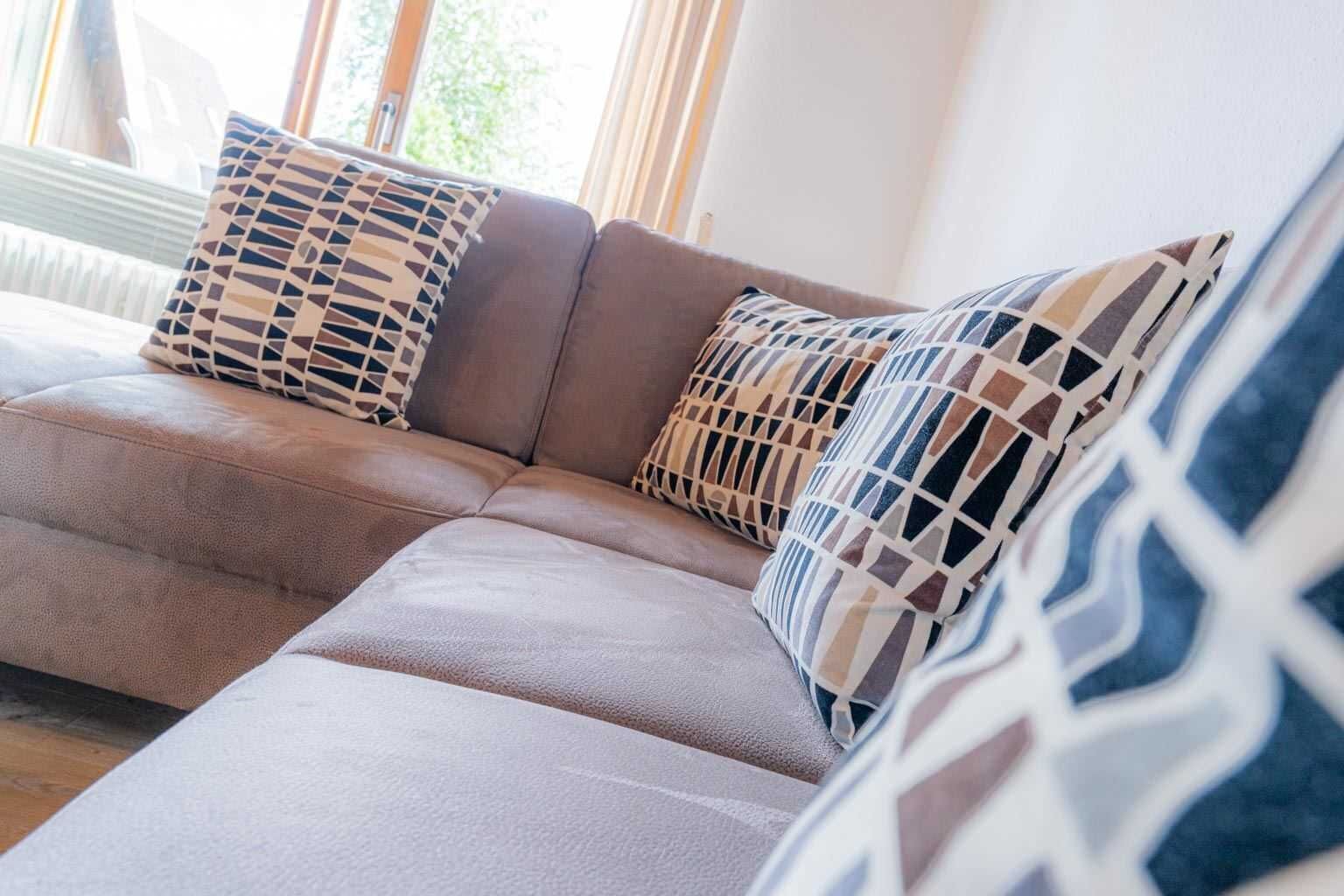 A beige sectional sofa with patterned cushions, positioned near a window with curtains.