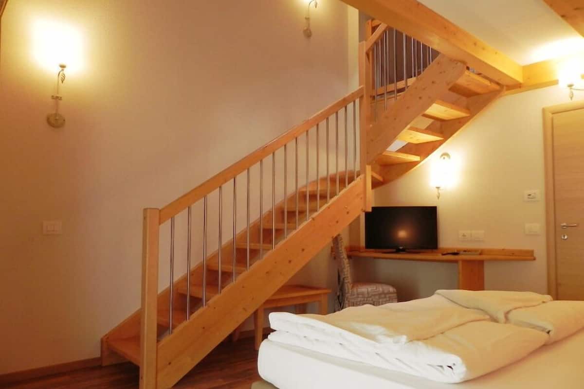 Une chambre avec un escalier en bois, un lit recouvert de draps blancs, un téléviseur posé sur un meuble en bois et une porte.