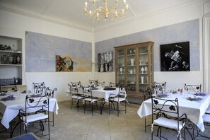 Une salle à manger avec des tables disposées pour accueillir quatre personnes, chacune étant recouverte d’une nappe blanche et entourée de chaises en métal noir. Un lustre est suspendu au-dessus des tables, et les murs sont ornés d’œuvres d’art.
